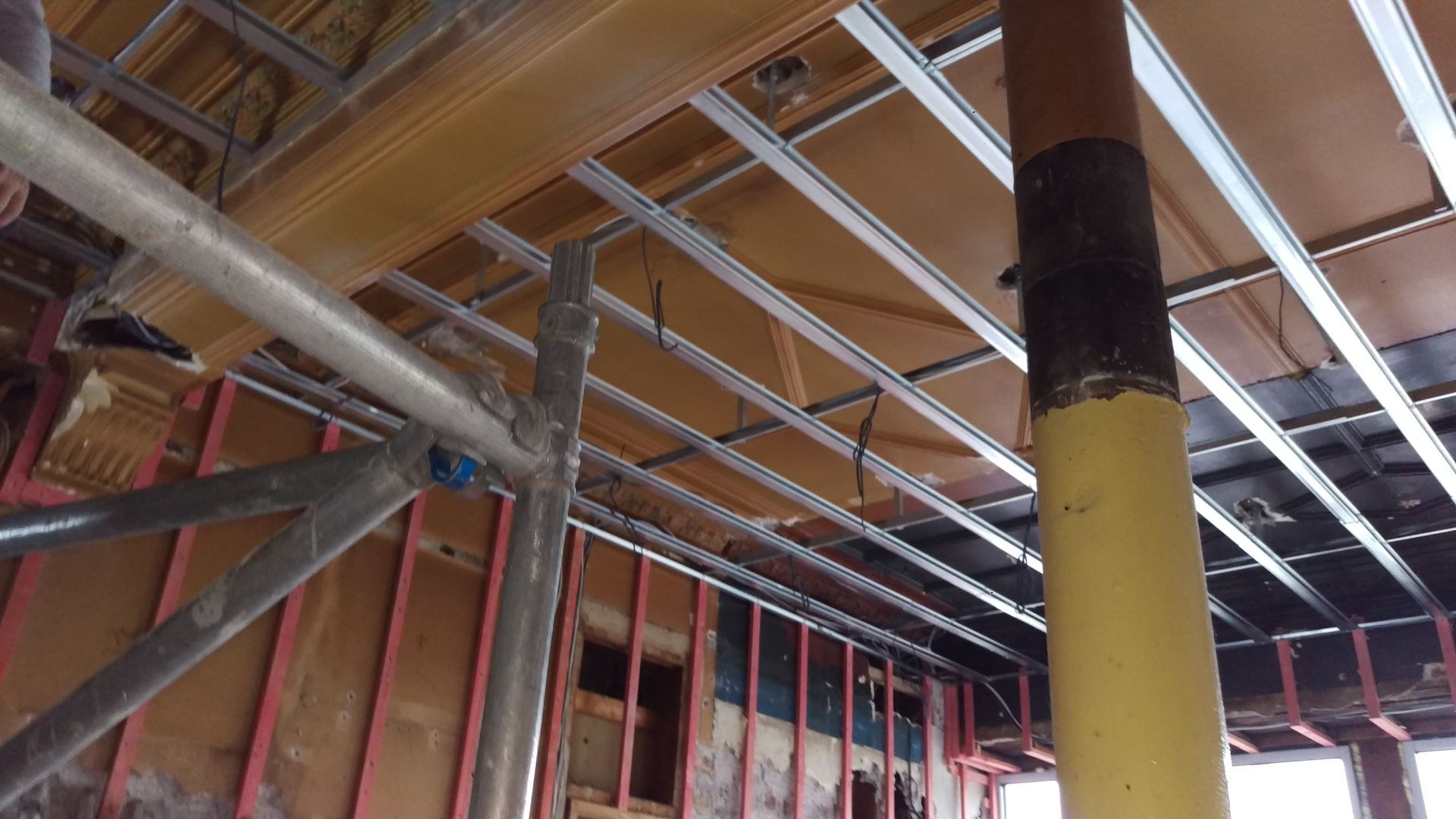 Interior construction: metal ceiling grid, wood paneling, scaffolding, and a yellow pipe.