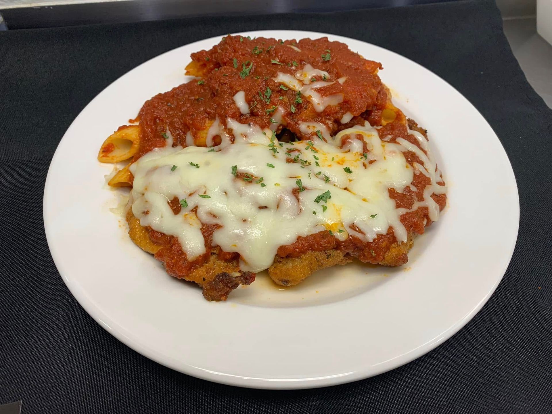 A breaded chicken parmigiana topped with melted mozzarella, marinara sauce, and herbs, served with penne pasta.