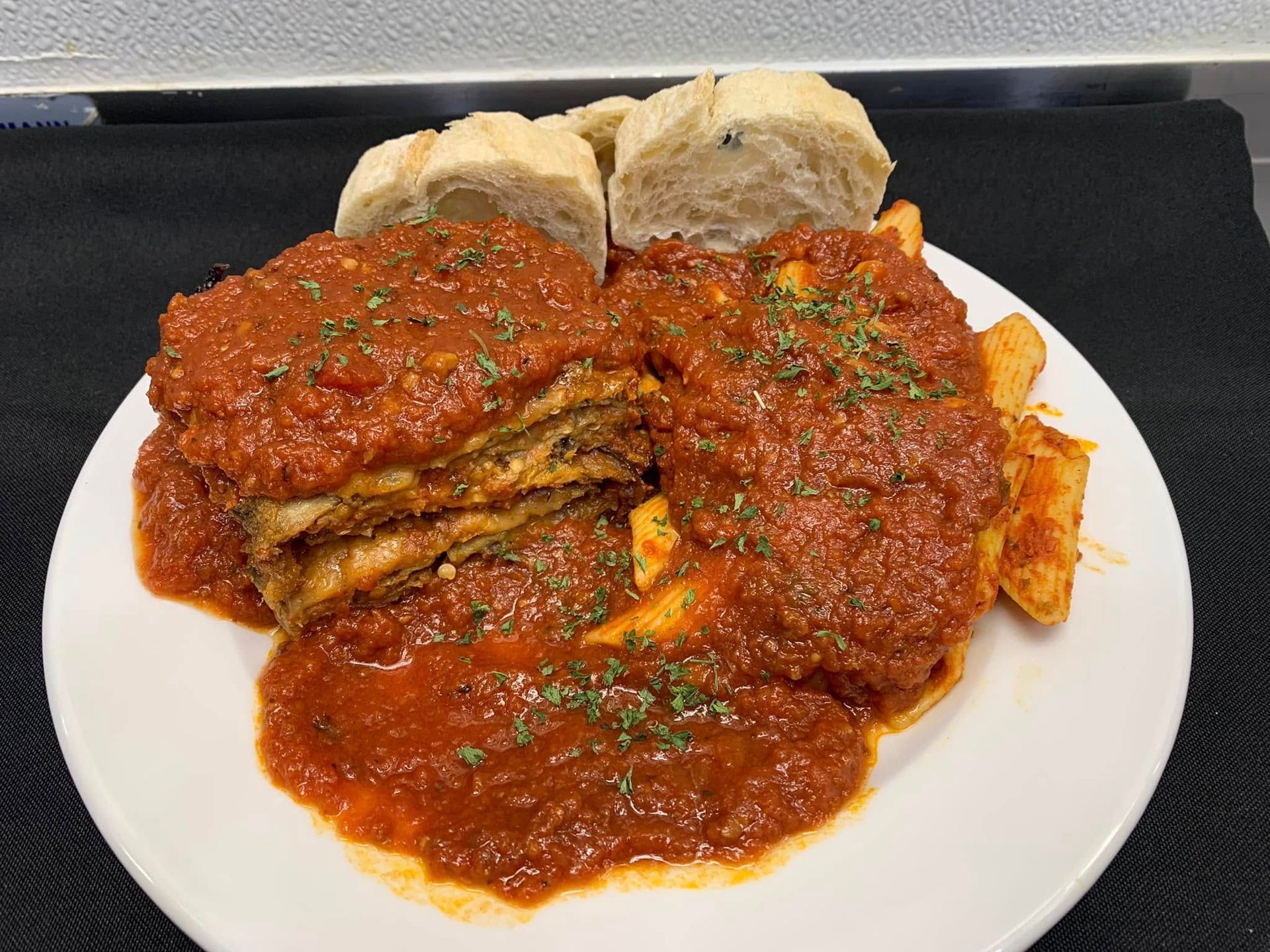 A plate of lasagna topped with red sauce and herbs, served with a side of fries and sliced bread on a black background.