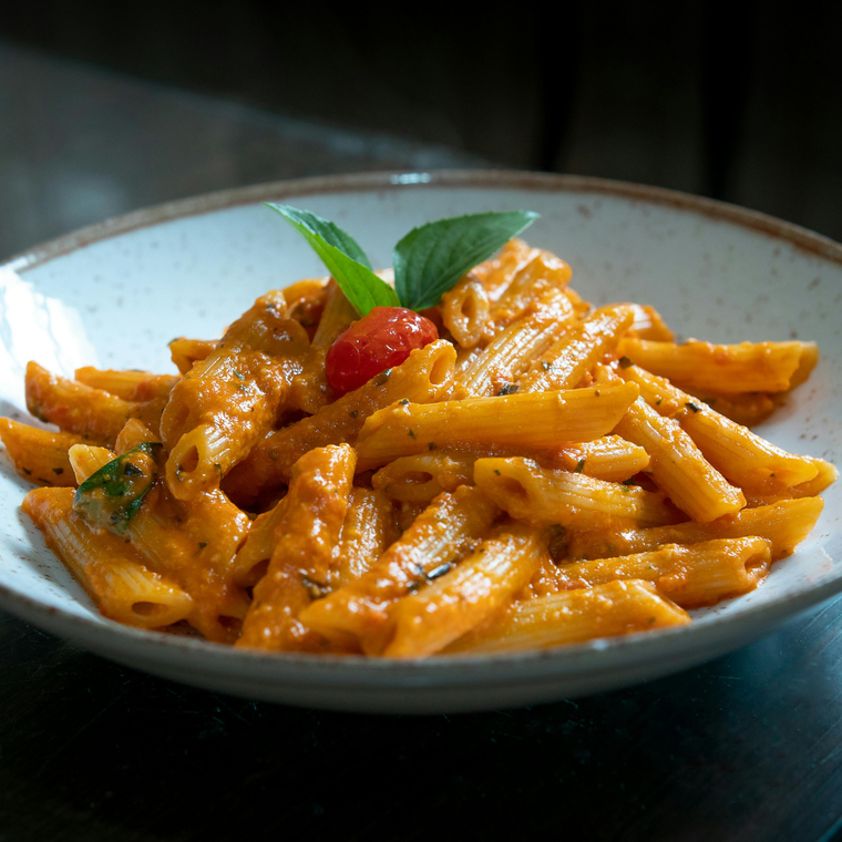 A bowl of penne pasta coated in a creamy orange sauce, topped with a cherry tomato and fresh basil leaves.