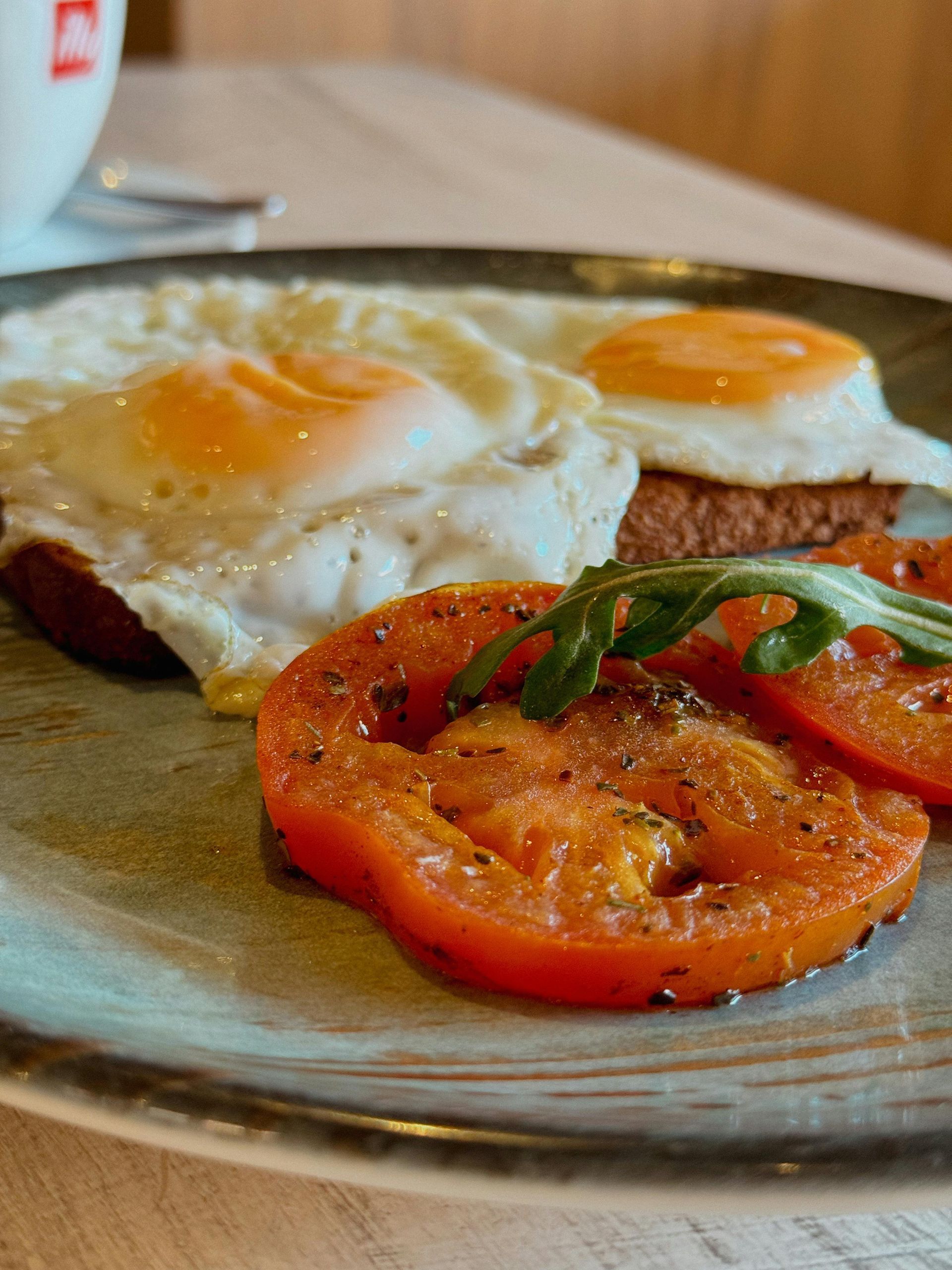 Two fried eggs on toast with seasoned tomato slices and a sprig of arugula on a ceramic plate.