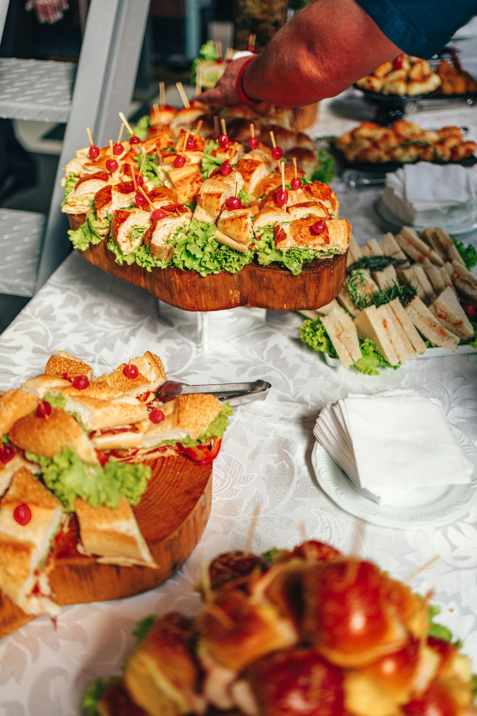 A buffet table featuring platters of sandwiches and bite-sized appetizers, with a hand reaching toward the food.