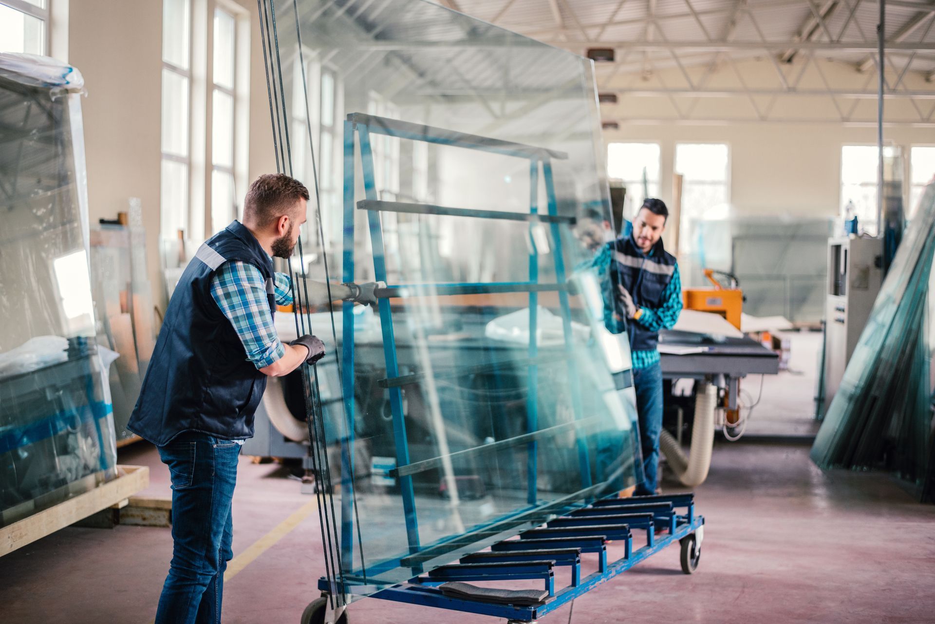 Two workers in a glass factory carefully maneuver a large glass panel on a wheeled rack.