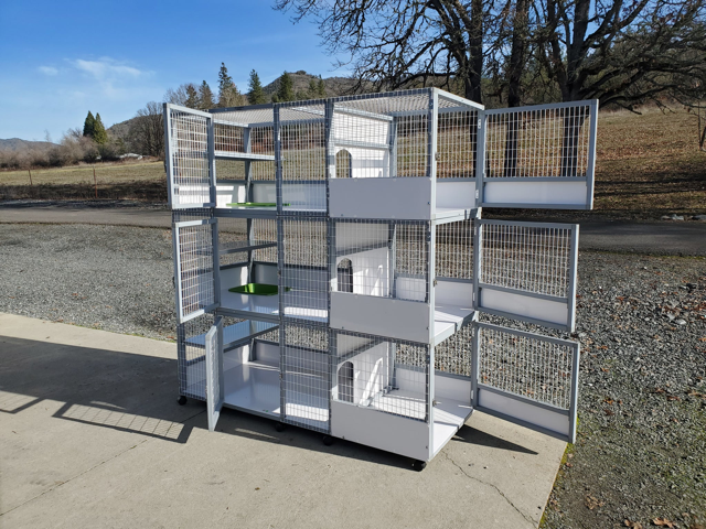 Gray cat cage with wire mesh sides, white shelves, and small entry holes. Wooden floor background.