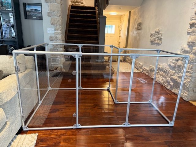 Clear pet enclosure on a wood floor, near a staircase and stone wall.