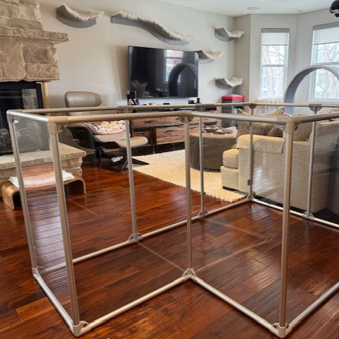 A white PVC pipe enclosure sits on a wood floor in a living room. Cat shelves are on the wall.