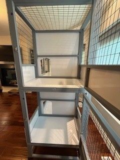Gray cat cage with wire mesh sides, white shelves, and small entry holes. Wooden floor background.