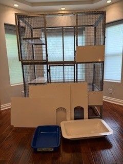 Cat cage with white panels and two litter boxes in a room with wood flooring and windows.