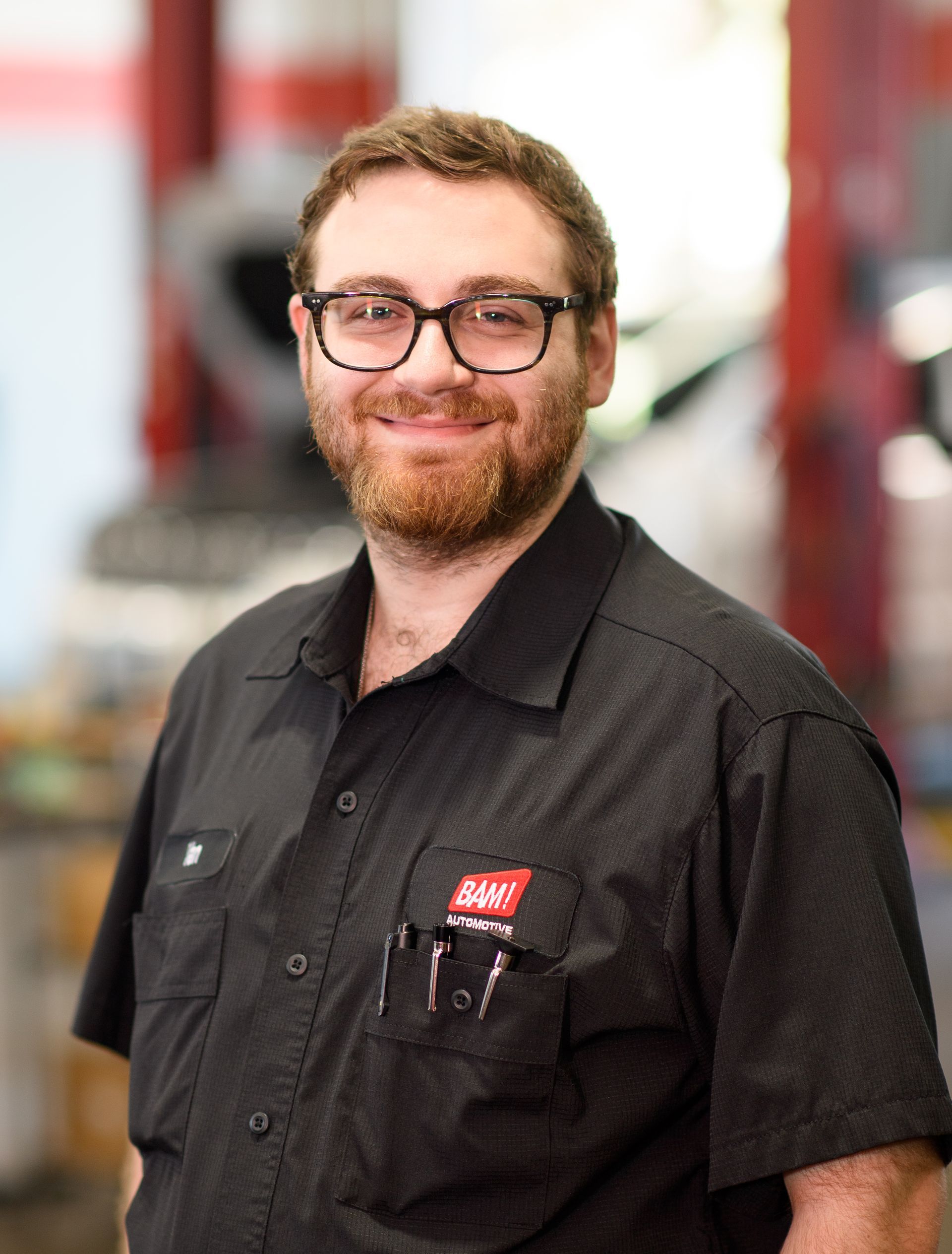 Man with glasses and beard, wearing a black work shirt. Smiling, standing in a workshop with machinery.