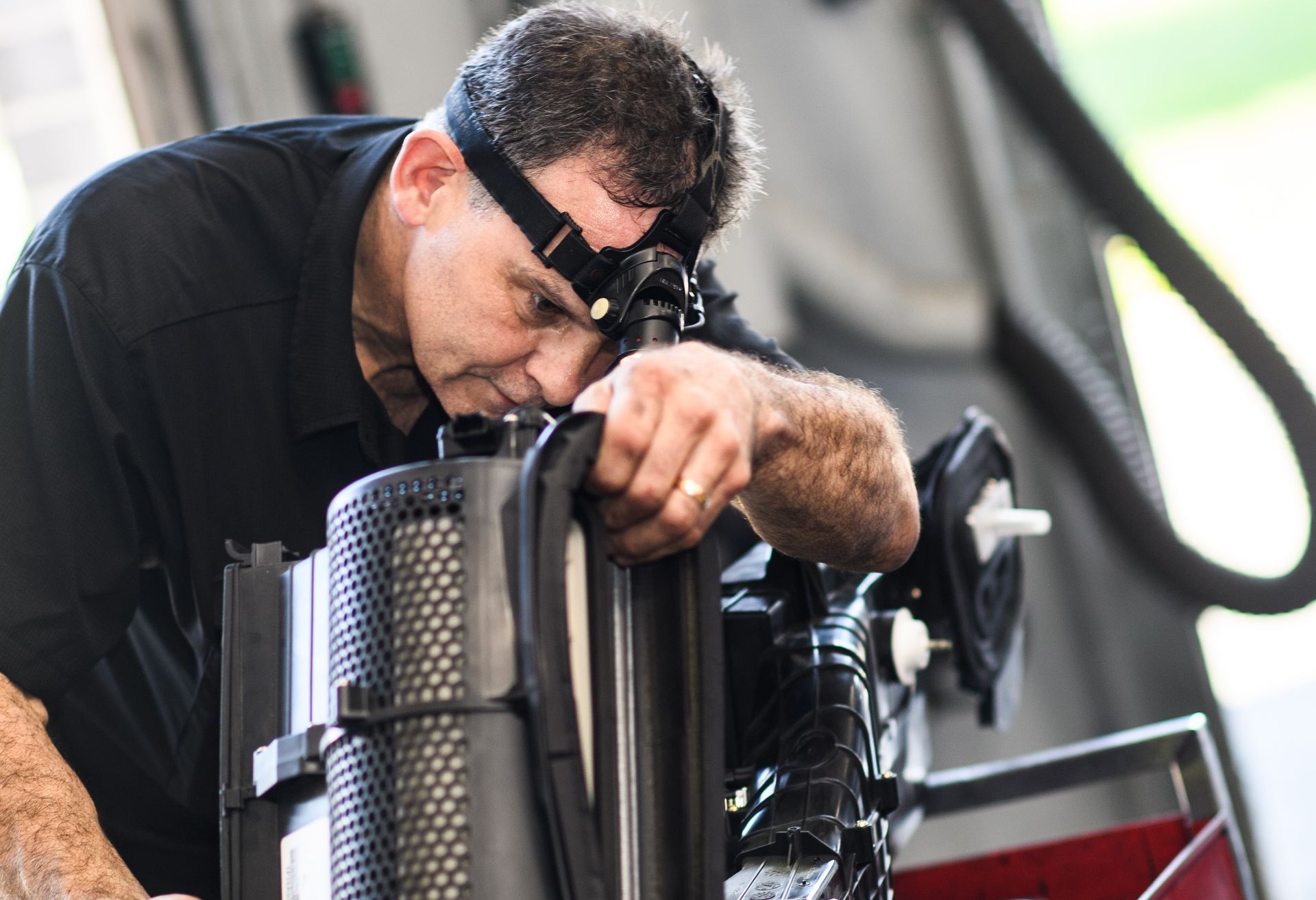 Man wearing headlamp, inspecting machinery. Black shirt, indoor setting.