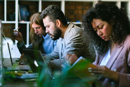 Three people working at desks, focused on laptops and notebooks in an office setting.