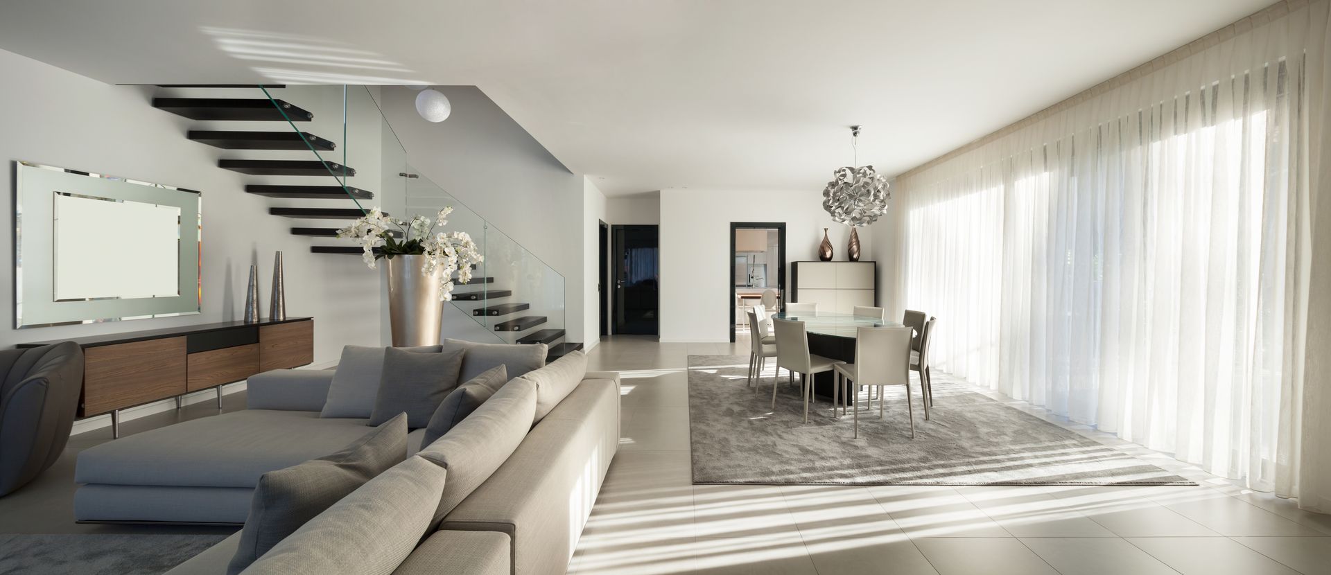 Bright modern open-plan living and dining room with gray sofa, staircase, and sunlit sheer curtains