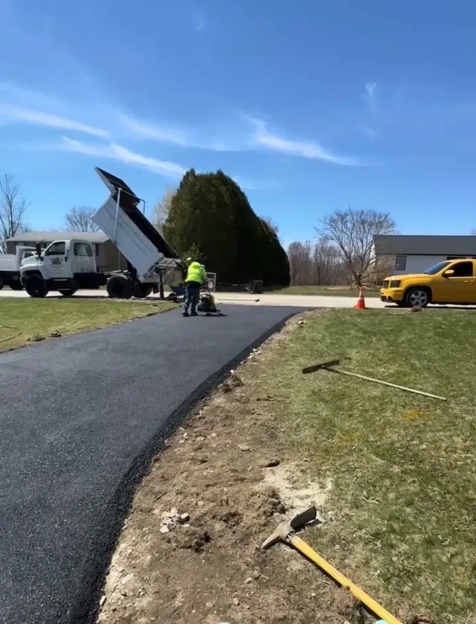 Paving crew laying fresh asphalt on a driveway, with a truck dumping material. Bright blue sky, sunny day.