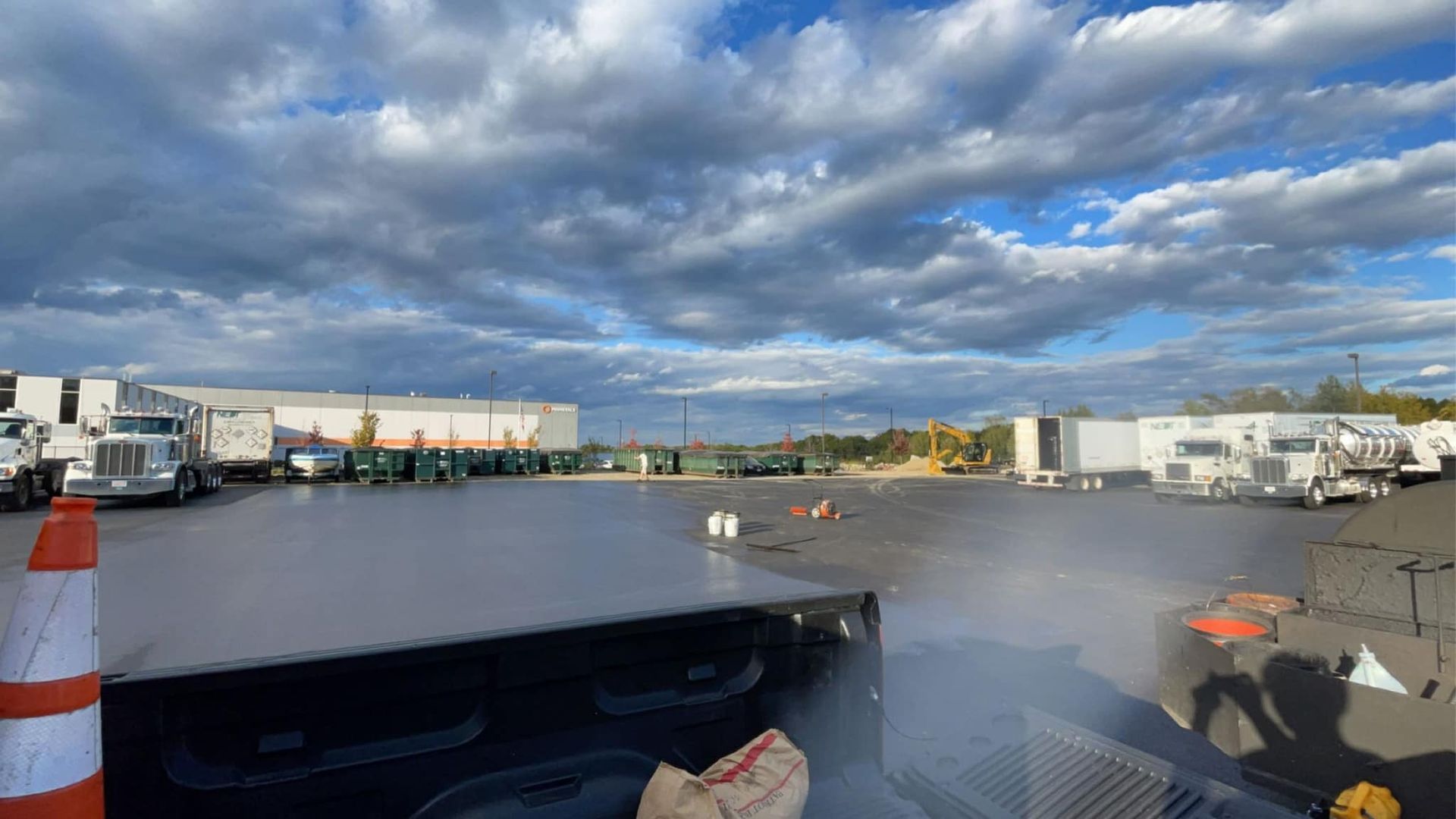 Trucks parked at a lot under a cloudy sky. Orange cones and possible fog or steam visible.