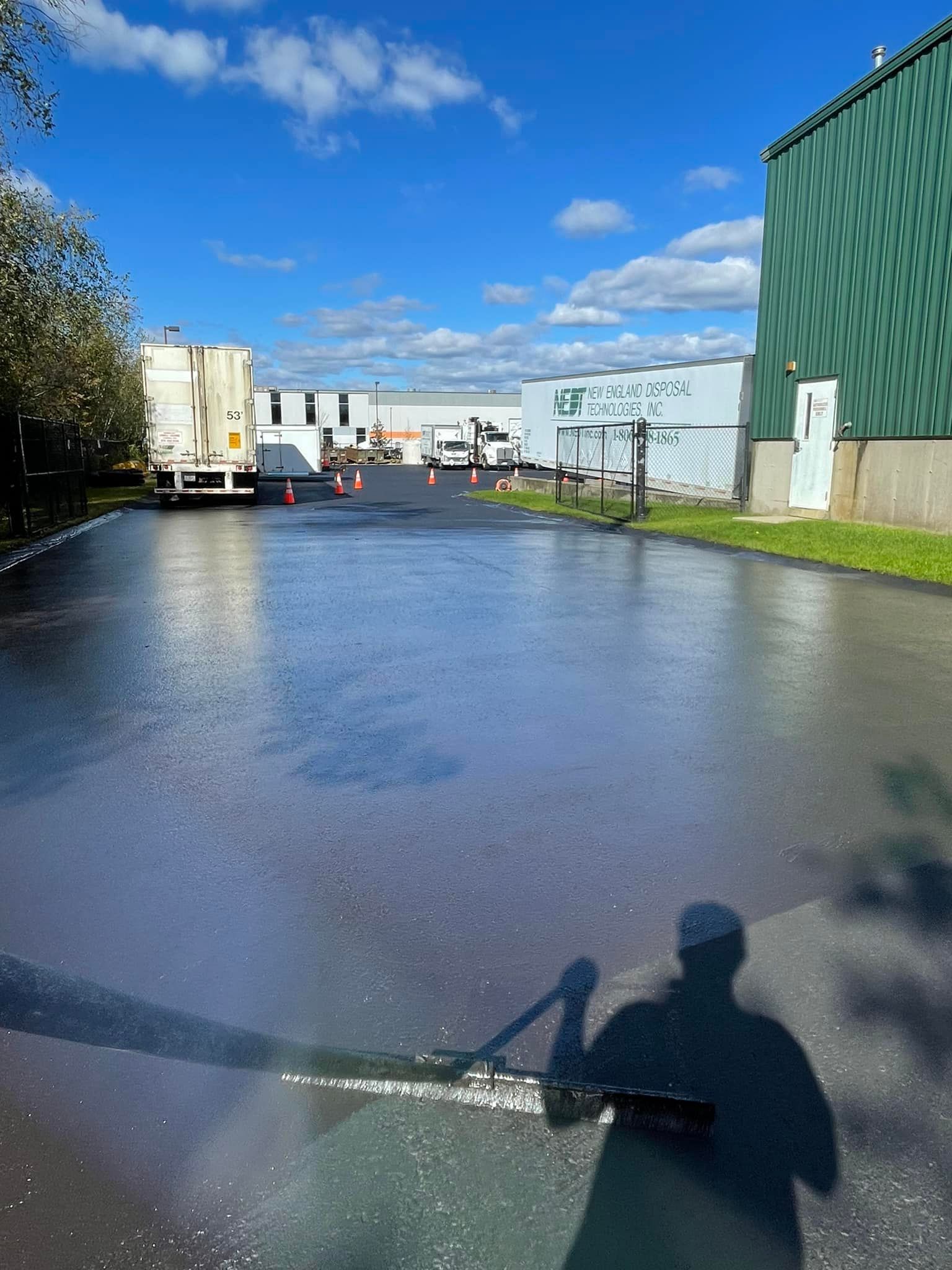 Person washing a wet asphalt parking lot with a long-handled squeegee on a sunny day.