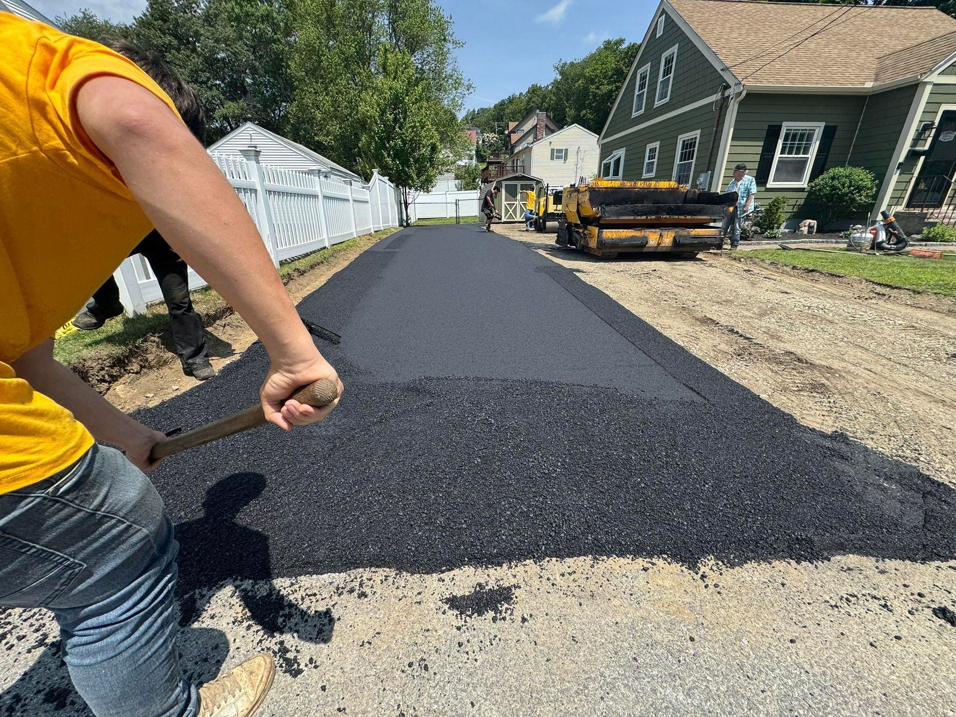 Person in yellow shirt shoveling asphalt on a freshly paved road in front of houses.