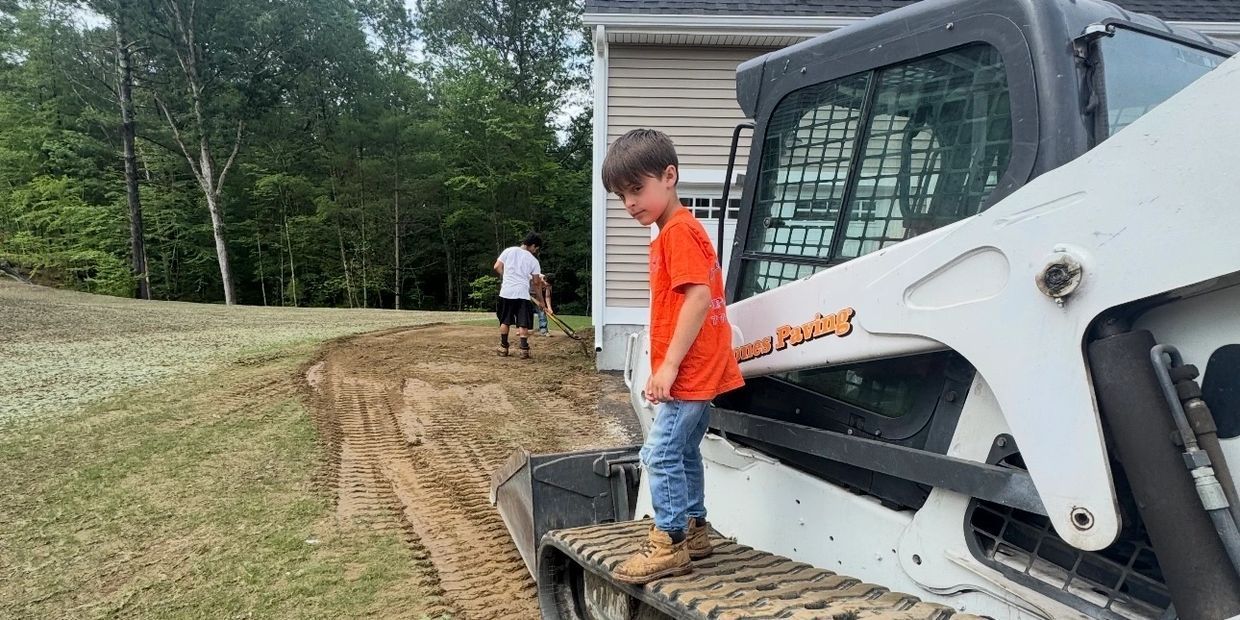 A young boy in an orange shirt stands on a skid steer, with a man working nearby.