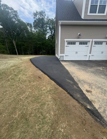 Asphalt driveway leading to a beige house with a two-car garage. Dry grass surrounds the driveway.