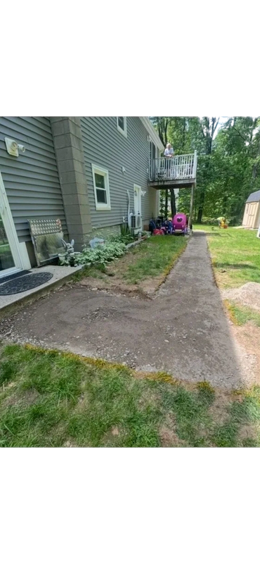 Gravel pathway alongside a house with a deck. Lush green grass, gray siding, and trees in the background.
