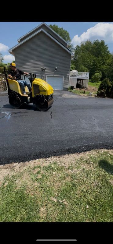 A yellow road roller compacting freshly laid asphalt driveway next to a grassy lawn.