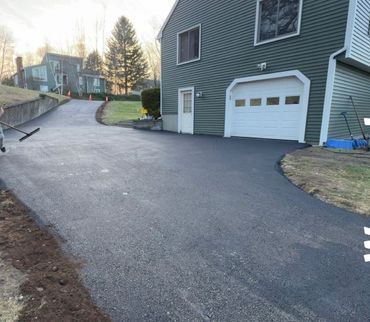 Freshly paved asphalt driveway leading to a two-story green house with a white garage door; worker raking gravel at the edge.