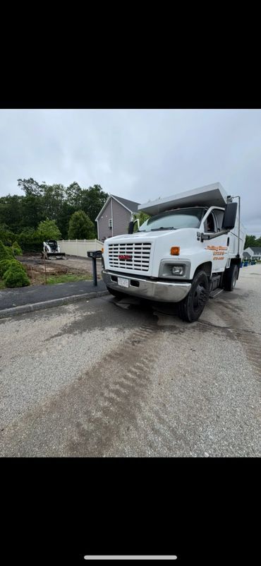 White dump truck on a gravel road in front of a house with a cloudy sky in the background.