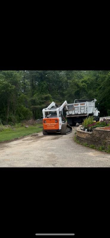 An orange and white bobcat loading a white dump truck on a gravel road surrounded by trees.