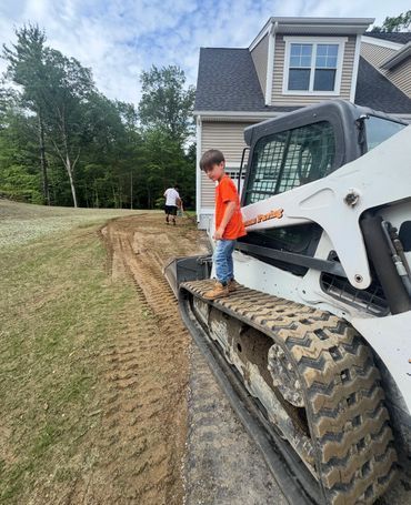 Boy standing on a skid steer tread. Man raking a path in background near a house.