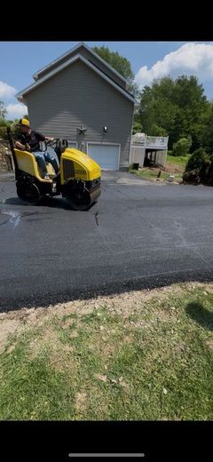 A worker on a yellow asphalt roller compacts a new black driveway, in front of a gray house.