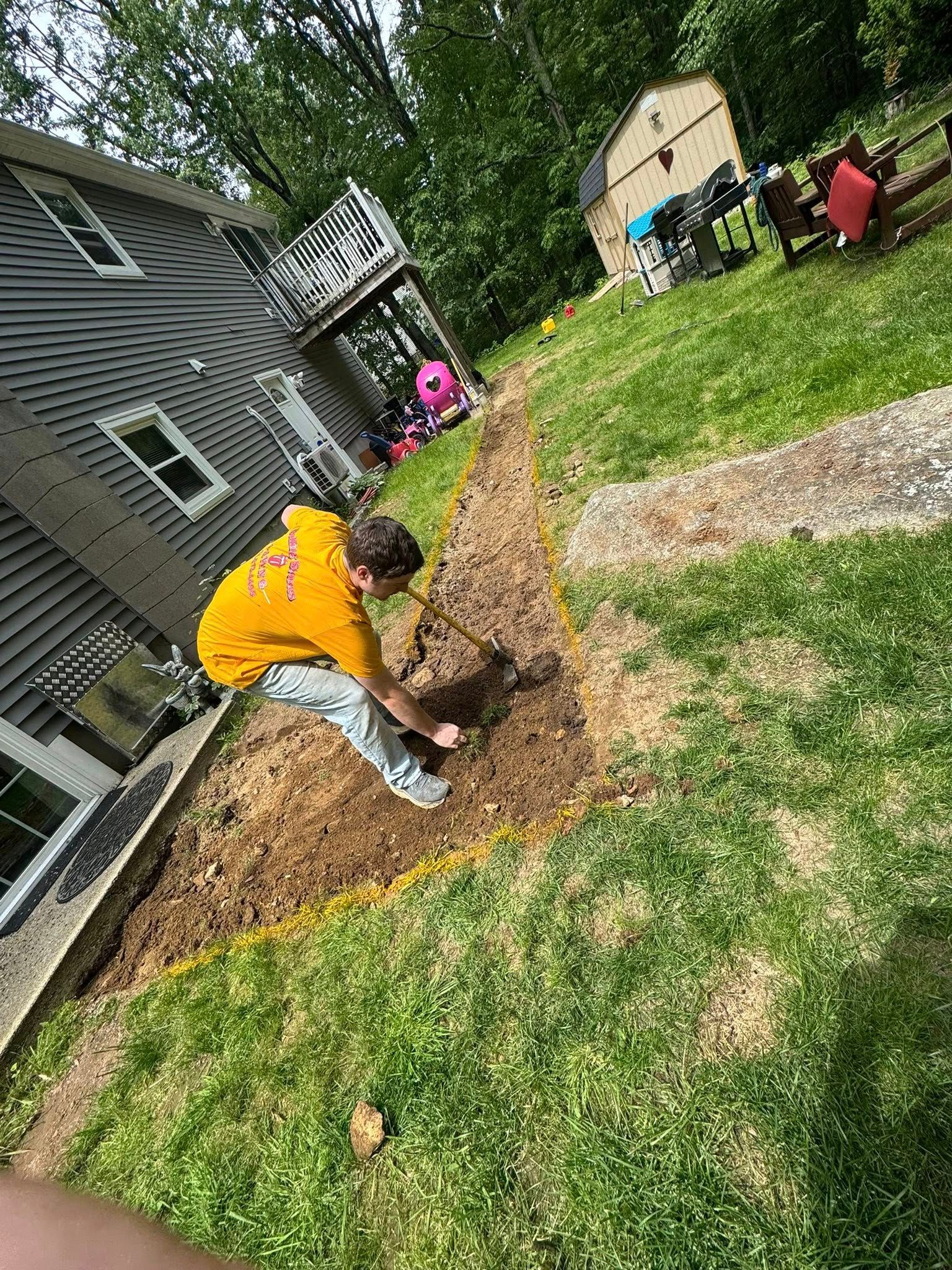A person in a yellow hoodie digs in a dirt patch next to a house.