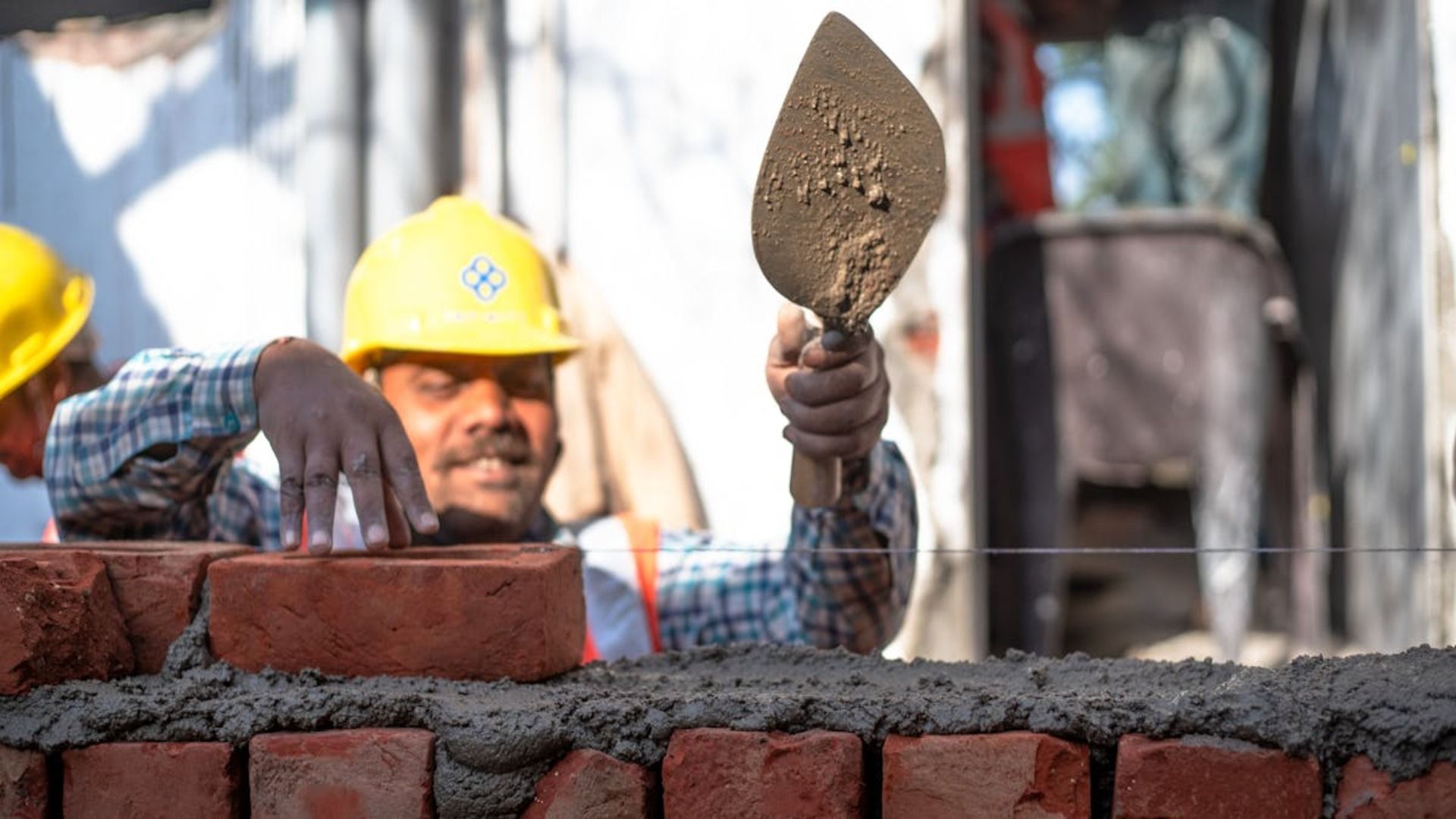 Albañil usando una paleta para aplicar mortero en una pared de ladrillos, usando un casco amarillo.