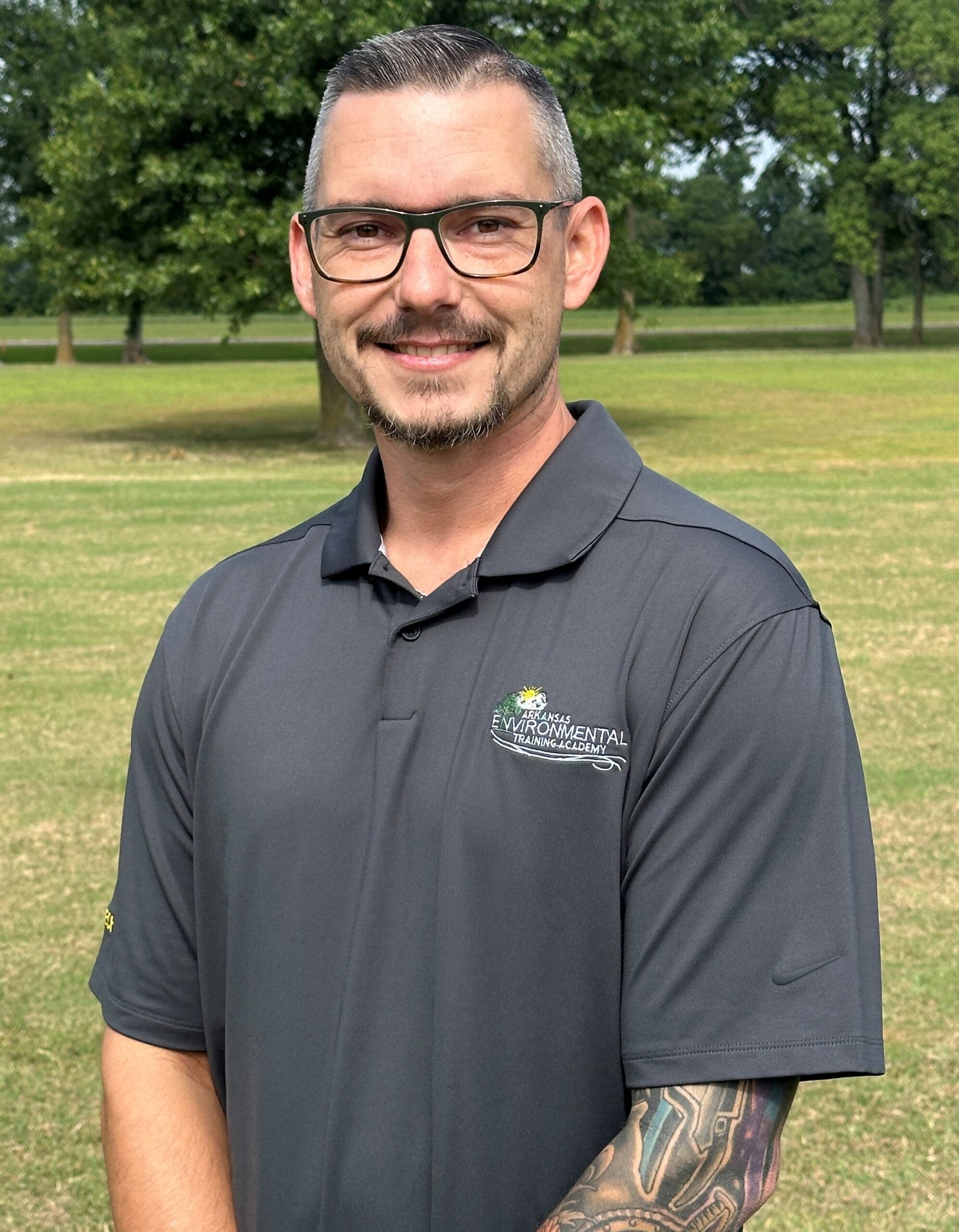 A man with glasses and a tattoo on his arm is standing in a field.