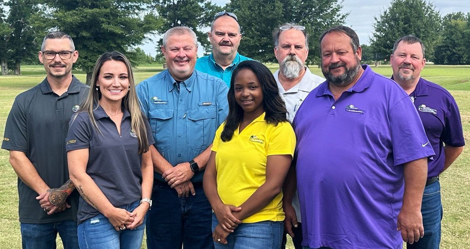 Group of nine people posing outdoors, smiling. They wear various colored polo shirts and stand in front of a green grassy area.