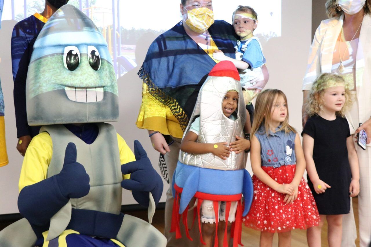 A group of children are standing around a mascot giving a thumbs up.