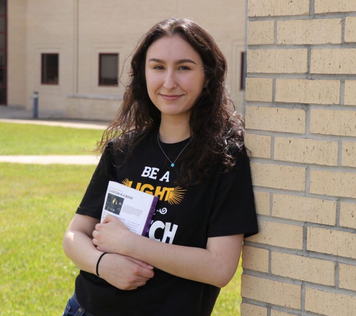 A woman wearing a black shirt that says be a light is leaning against a brick wall