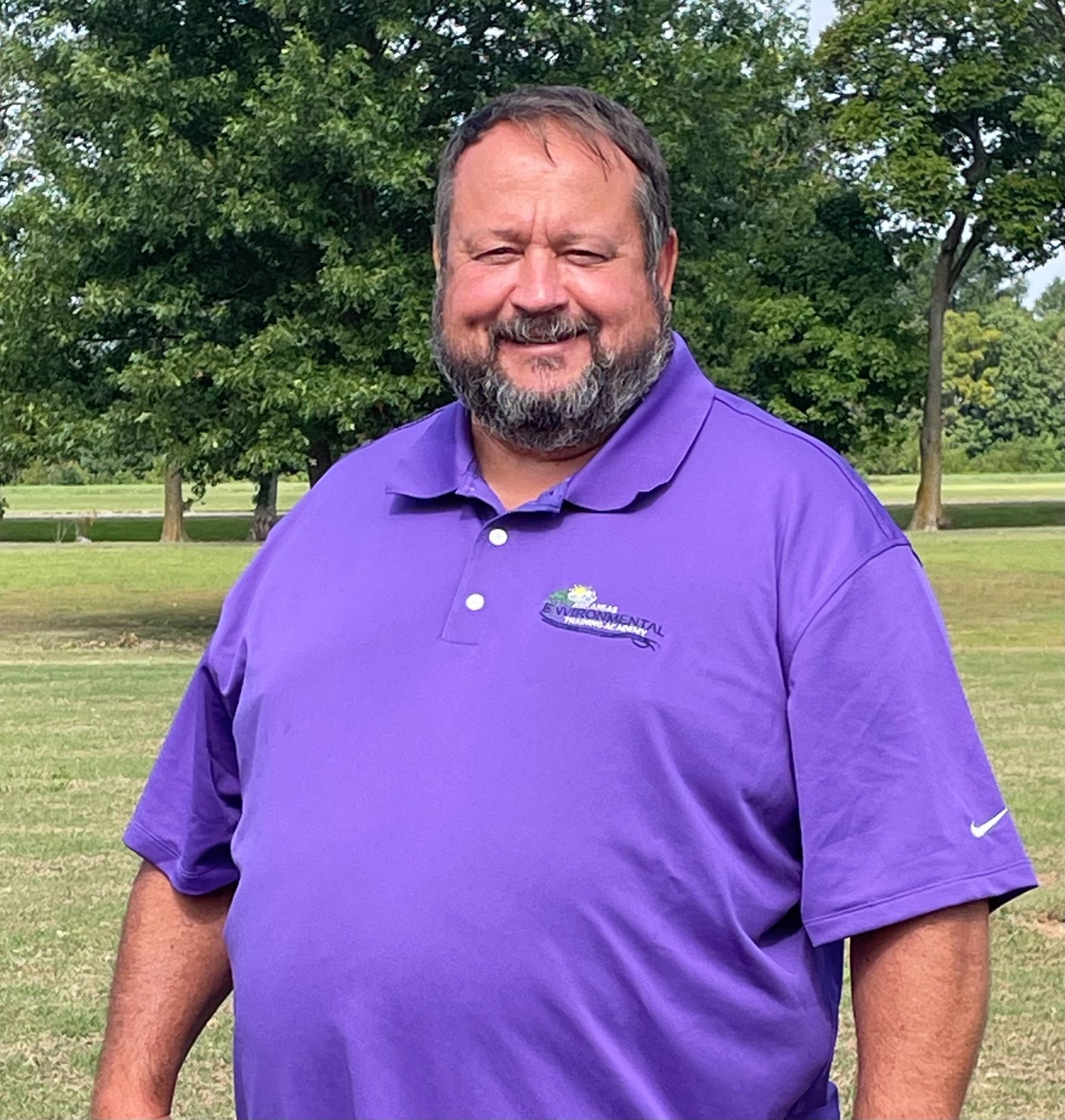 A man with a beard wearing a purple shirt is standing in a field.