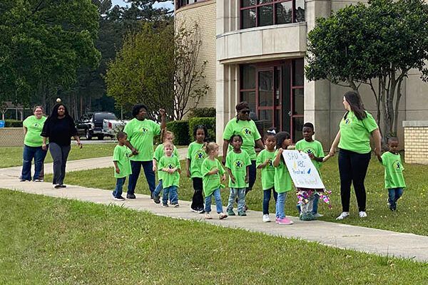 A group of children wearing green shirts are walking down a sidewalk.