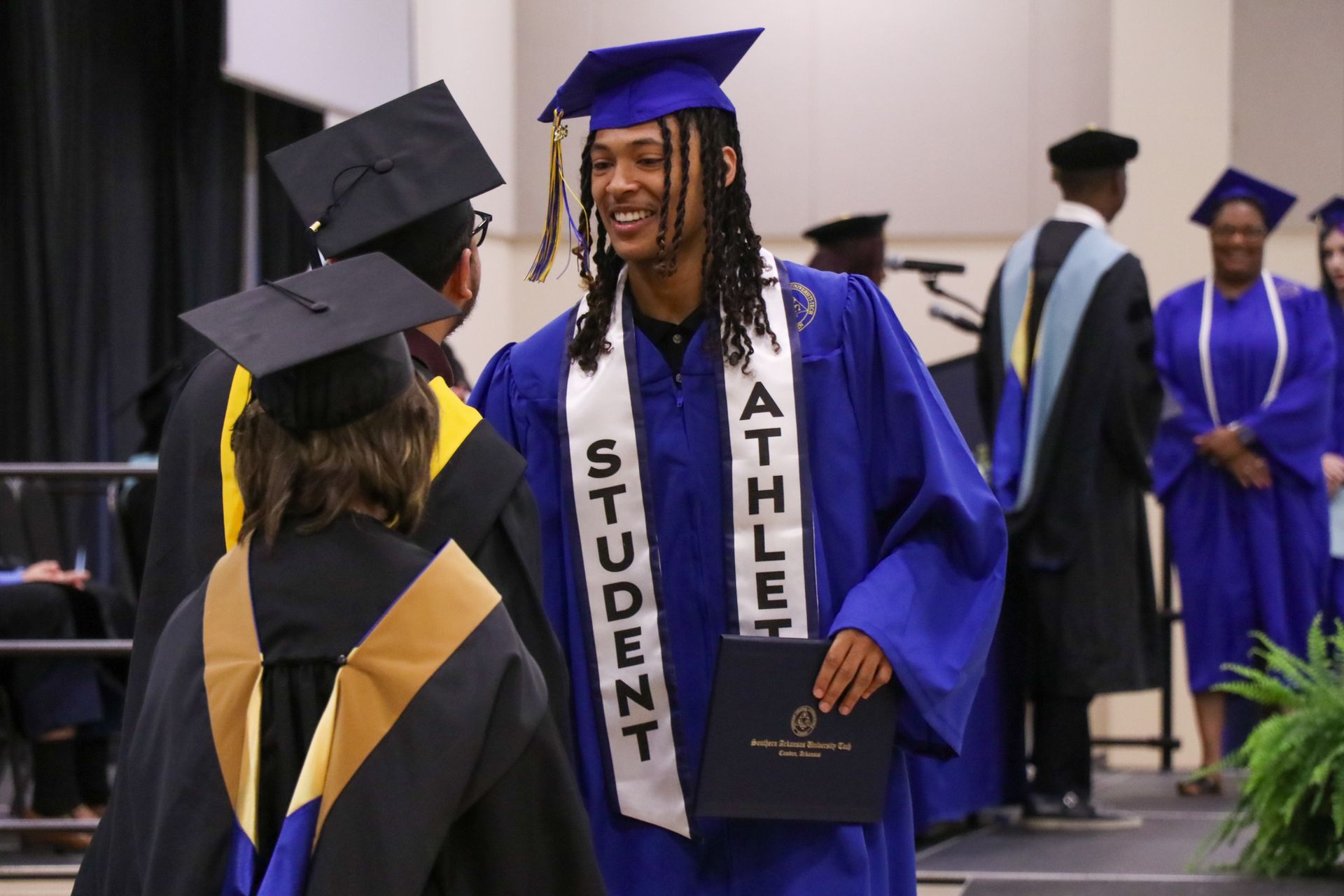 Graduating student athlete in blue gown smiles and holds diploma.