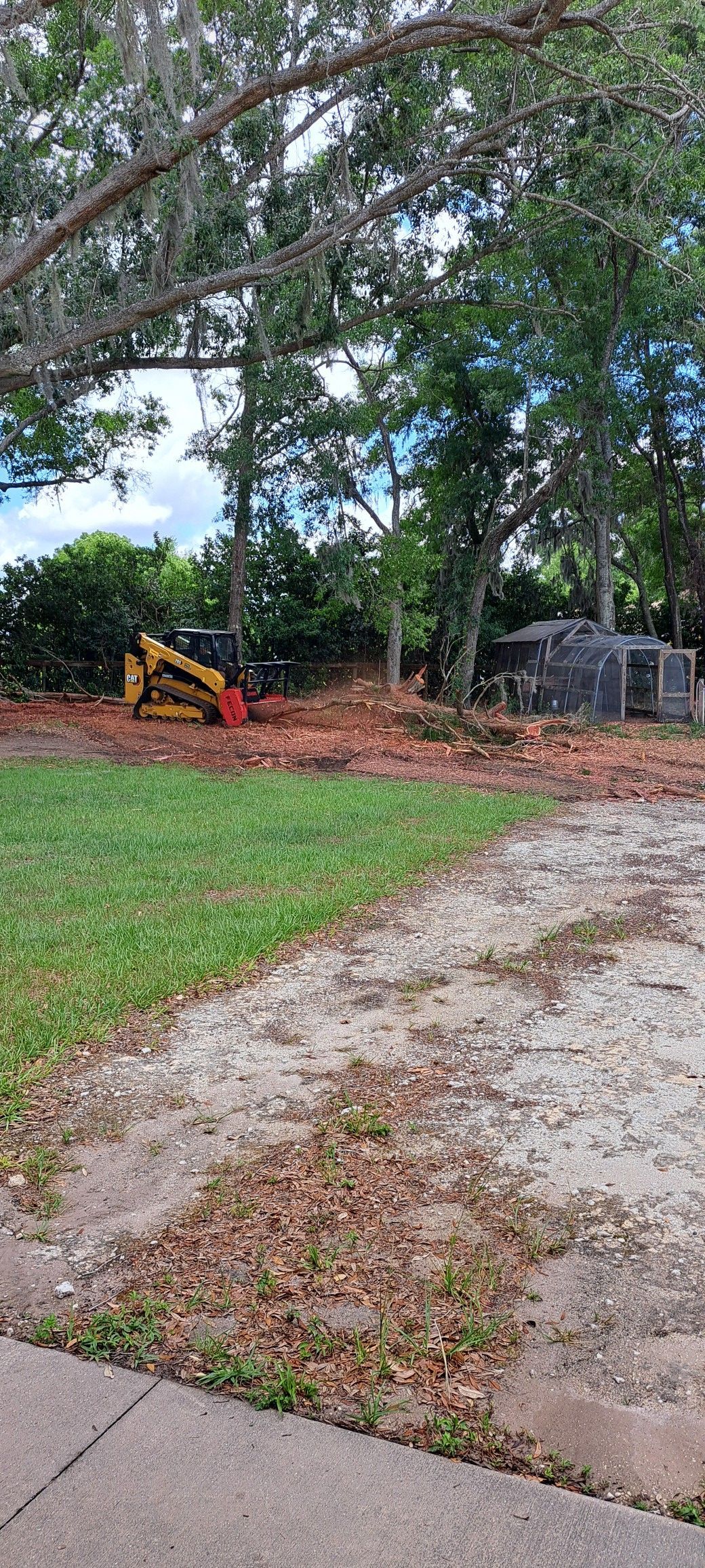 A yellow skid steer tractor clears debris behind a green lawn and gravel driveway under trees.