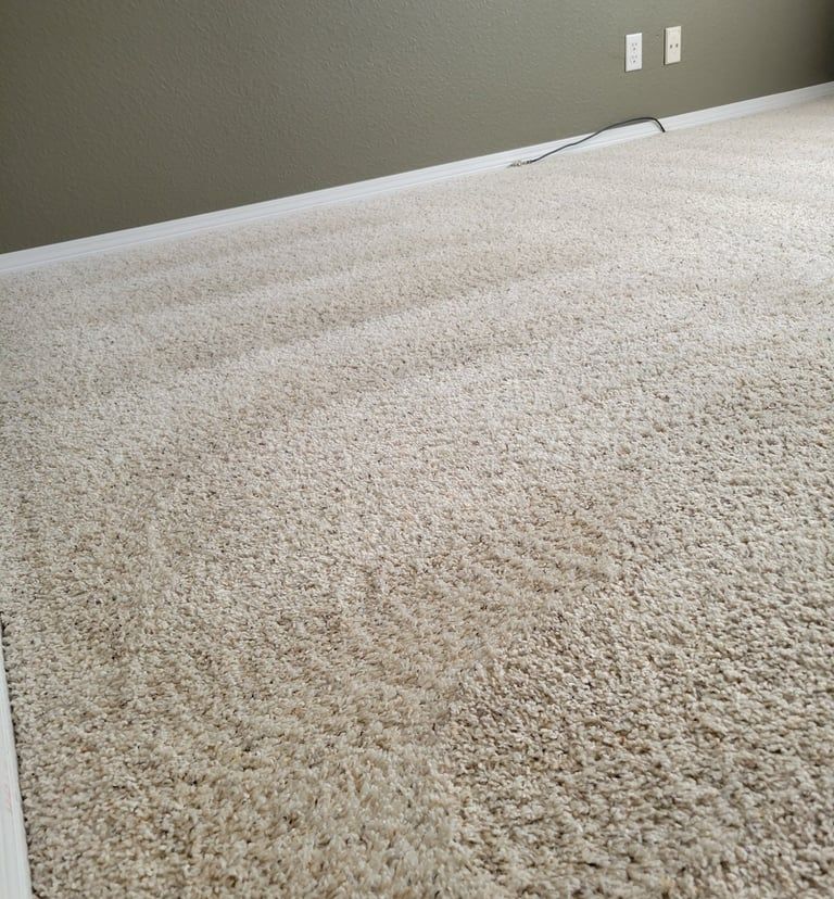 A close-up of light beige carpet with visible vacuum lines, meeting a white baseboard and a grey wall.