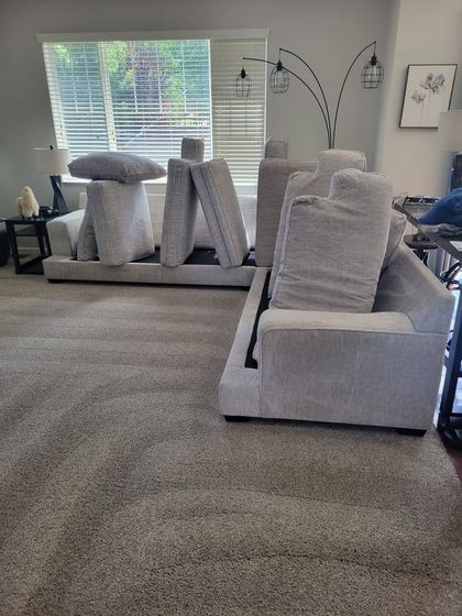 A light-gray sectional sofa with detached back cushions propped upright on the seats in a carpeted living room.