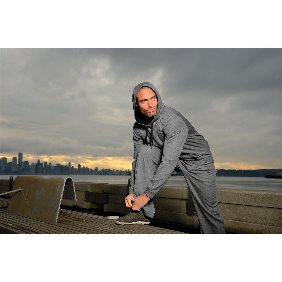 A man in a hoodie is tying his shoes on a pier.