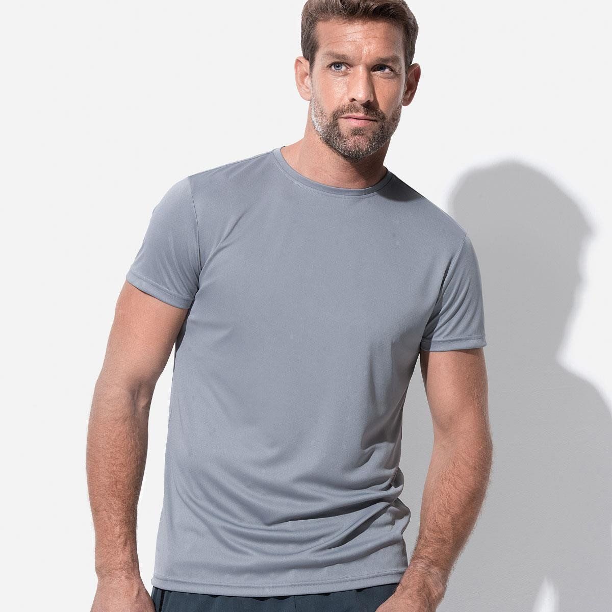 A man wearing a grey t-shirt is standing in front of a white wall.