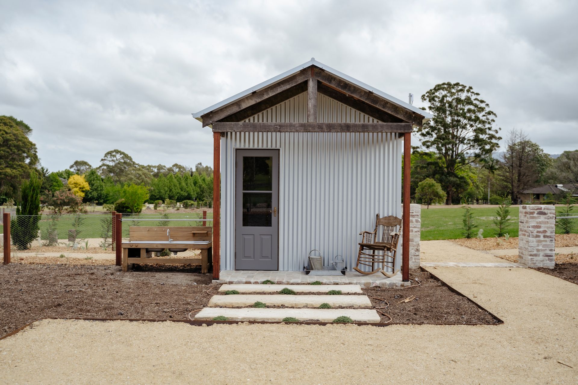 Small, corrugated metal shed with a gray door, a bench, and rocking chair outside, surrounded by gravel path  — Stark Building & Constructions In Shoalhaven, NSW