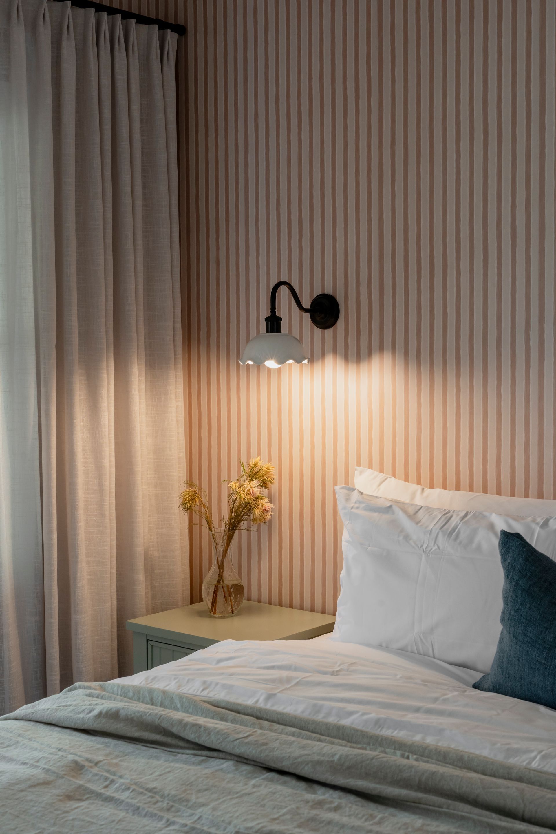Bedroom with a bed, nightstand, lamp, and floral arrangement. Pink striped wallpaper, gray curtains, and a teal pillow  — Stark Building & Constructions In Berry, NSW