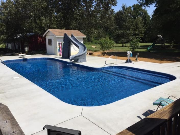 A deep blue, irregularly shaped swimming pool with a grey slide and concrete deck, set in a sunny backyard.
