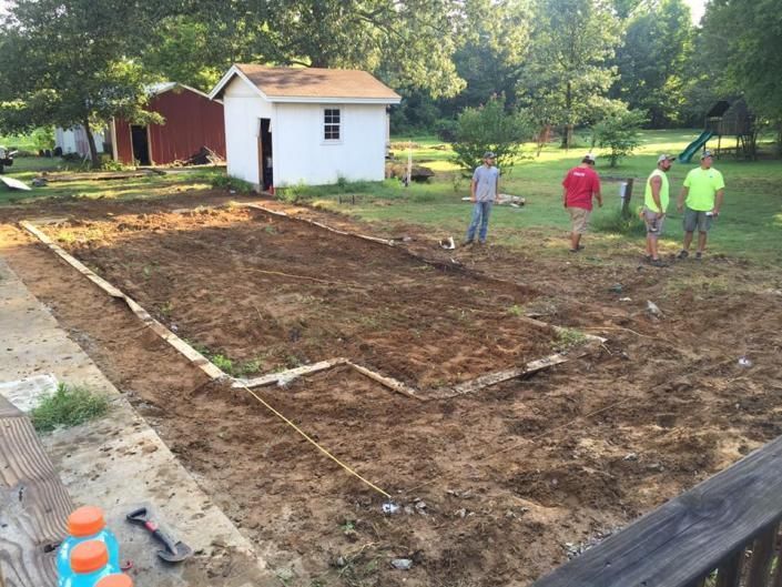 Four people stand near a rectangular excavation site in a grassy backyard with a white shed in the background.