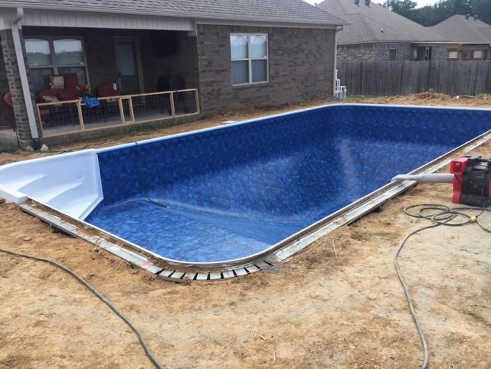 An empty rectangular swimming pool with blue patterned liner and white steps, set in a backyard under construction.