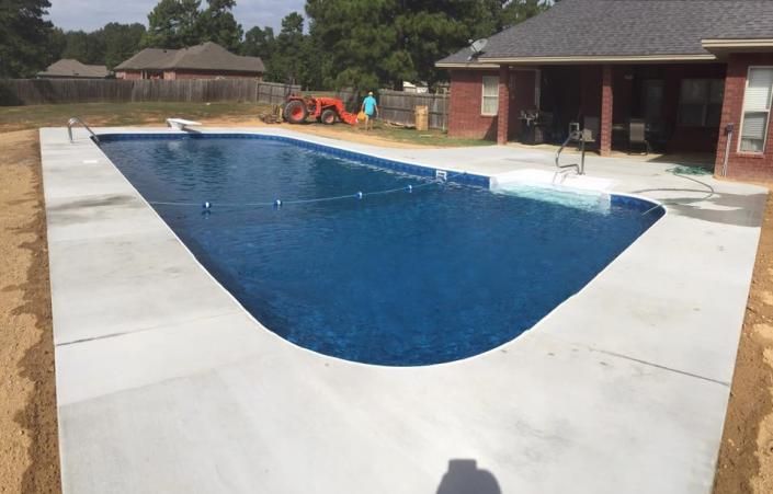 A newly installed rectangular swimming pool with blue water and surrounding concrete decking near a brick house.