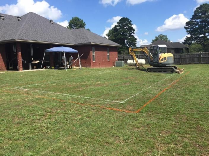 A construction site in a backyard with white and orange spray-painted markings, a house, and an excavator.
